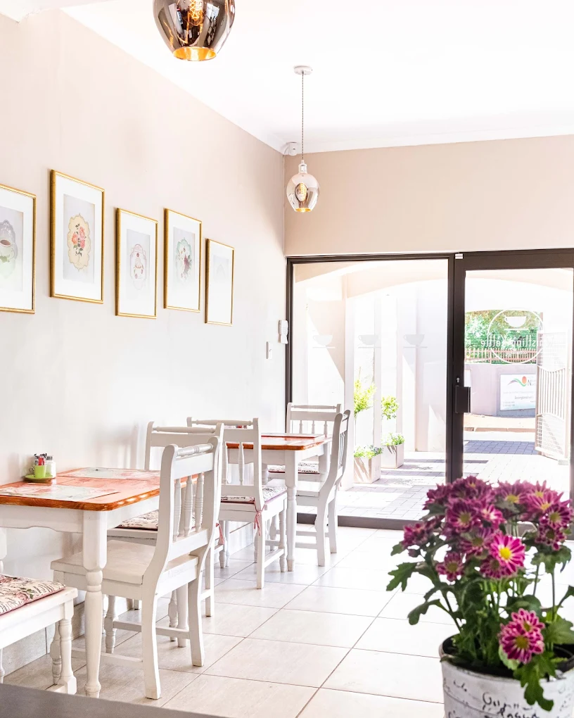 Inside Whistling Kettle — sunlit dining room with white furniture, framed botanical prints and pink florals